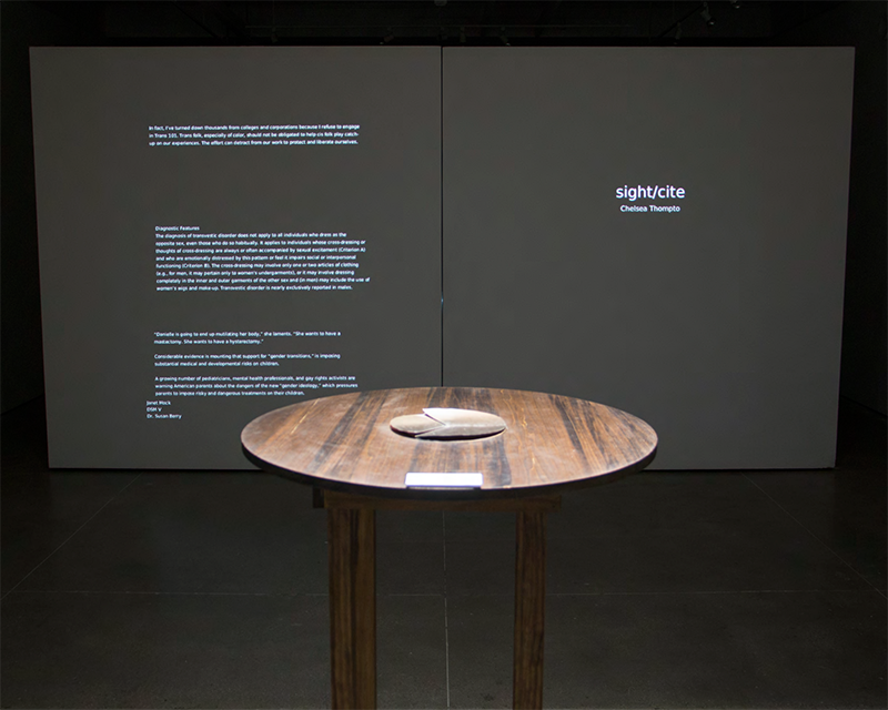 A four legged table with a circluar top and circuler book stands in front of a wall featuring projected text.
